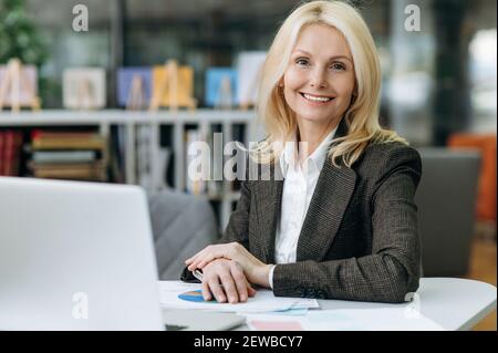 Fröhliche Mitarbeiterin sitzt am Schreibtisch im Büro und arbeitet mit Dokumenten. Elegante reife Geschäftsfrau in stilvollem Anzug schaut auf die Kamera, lächelnd Stockfoto