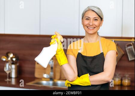Zufrieden lächelnd reife asiatische Hausfrau oder Putzfrau in gelben Handschuhen und einer Schürze, hält Waschmittel, bereit zu Hause oder im Büro Reinigung, schaut auf die Kamera Stockfoto