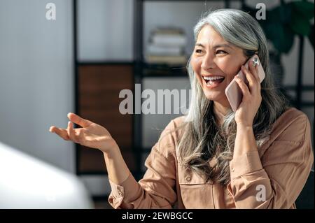 Zufriedene, erfolgreiche, grauhaarige Dame mittleren Alters, Geschäftsfrau, haben Telefongespräch mit Kollegen oder Freund, gestikulieren mit der Hand, schaut weg und lächeln glücklich Stockfoto
