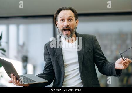 Ein männlicher Angestellter mittleren Alters überraschte mit unerwarteten Nachrichten und schaute auf die Kamera. Schockiert Geschäftsmann halten einen Laptop, bekommen Job-Förderung oder erhalten großen Gewinn aus erfolgreichen Projekt Stockfoto
