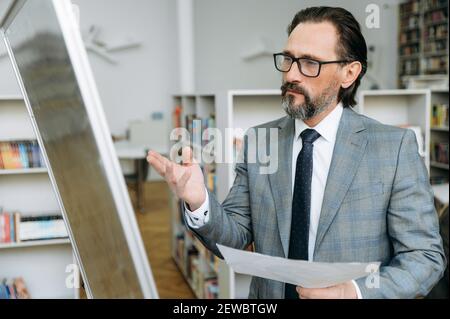 Konzentrierter reifer männlicher Mitarbeiter in Augenlaserinnen arbeitet an ernsten Projekt. Einflussreicher Geschäftsmann schaut nachdenklich auf das Flipchart, der im Büro steht Stockfoto
