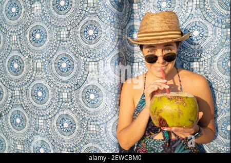 Eine attraktive Frau in Badeanzug, Sonnenbrille und Hut, die einen frischen Kokosnusscocktail trinkt Stockfoto