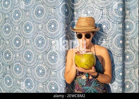 Eine attraktive Frau in Badeanzug, Sonnenbrille und Hut, die einen frischen Kokosnusscocktail trinkt Stockfoto