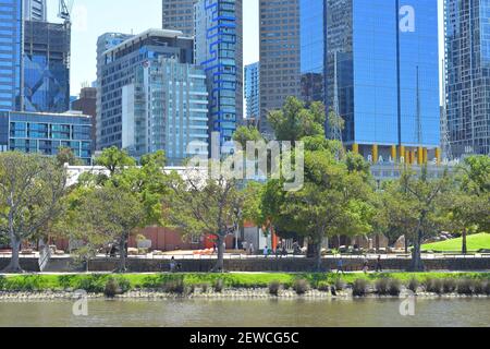 Fußweg mit gelegentlichen Schatten von Bäumen entlang des Flusses Yarra in Melbourne mit blauen Wolkenkratzern im Hintergrund. Stockfoto