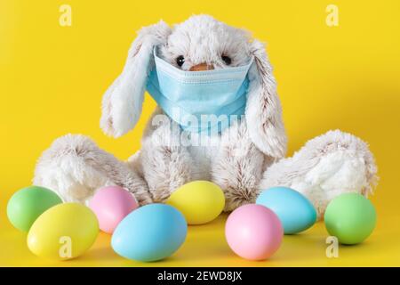 Ostern Stofftier Hase in Schutzmaske und farbige Eier auf gelbem Hintergrund. Ester Urlaub während Coronavirus Pandemiekonzept. Stockfoto