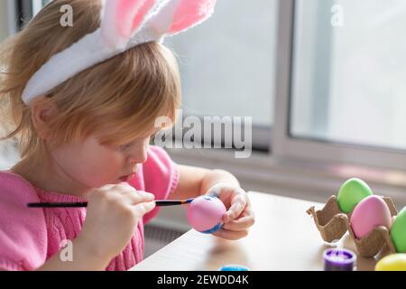 Kleines Mädchen in Osterhasen Ohren malen farbige Eier. Ostern Familienfeiertag zu Hause und Handwerk Konzept. Stockfoto