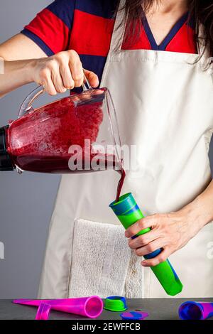 Eine kaukasische Mutter trägt Küchenschürze gießt Beerenpüree, das sie in ihrem Mixer in popscile Formen zum Einfrieren gemacht hat. Dies ist alles natürliche Bio-fr Stockfoto