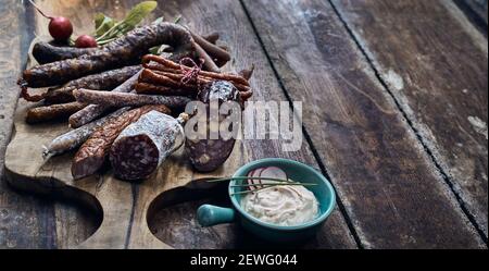 Hoher Winkel von appetitlich geräucherte und geräucherte Würste mit ganzen Radieschen in der Nähe Schüssel mit cremigem Käse auf Holztisch Stockfoto