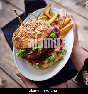 Hoher Winkel der Ernte anonyme Person zeigt Teller mit lecker burger mit frischem Gemüse und gebackenen Kartoffelscheiben Stockfoto