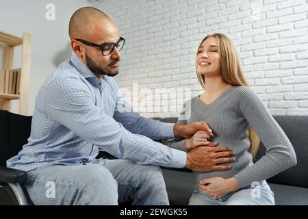 Behinderter afroamerikanischer Mann, der den Bauch seiner Schwangeren berührt kaukasische Frau, während sie auf dem Sofa zu Hause entspannen Stockfoto