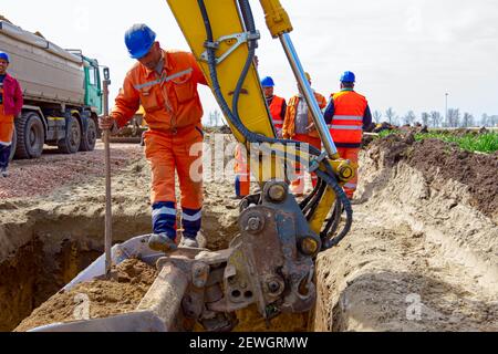 Arbeiter ist Sprung aus quadratischen Graben mit wenig Hilfe von Bagger Eimer voll von ausgegrabenen Boden. Stockfoto