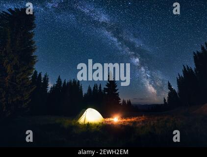 Panoramablick auf Nachtcamping im Tal mit großen Pinien. Brennendes Lagerfeuer und beleuchtetes Touristenzelt unter hellem Sternenhimmel mit Milchstraße am Abend. Tourismus in den Bergen Stockfoto