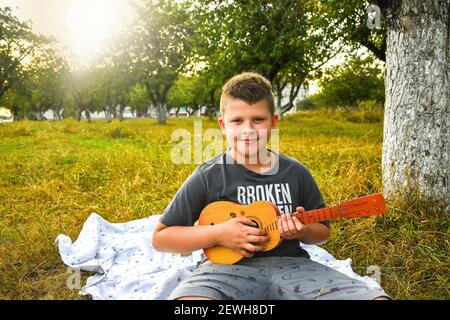 Kleiner Junge Gitarrist im Freien. Junge auf Stadtpark Sommerwiese genießen Tag Gitarre spielen. Freizeitaktivitäten im Freien. Stockfoto