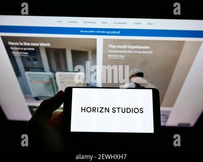 Person, die Mobiltelefon mit Firmenlogo des deutschen Gepäckherstellers Horizn Studios auf dem Bildschirm vor der Webseite hält. Konzentrieren Sie sich auf die Telefonanzeige. Stockfoto