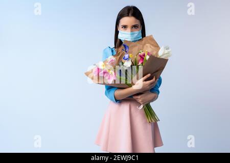 Porträt eines kaukasischen Mädchens in einer medizinischen Maske mit einem großen Strauß verwelkter bunten Tulpen in den Händen. Urlaubskonzept während einer Pandemie Stockfoto