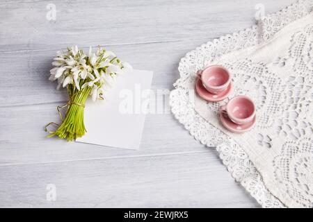 Bündel Galanthus mit einem leeren Stück Papier, flach liegend, Draufsicht. Zwei pinke Teetassen aus Keramik auf einem bestickten Stück Stoff Stockfoto