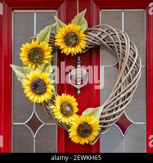 Ein Stock und Sonnenblume Herz geformte Tür Dekoration hängt an einer leuchtend roten und Glastür mit einem silbernen Klopfer. Stockfoto