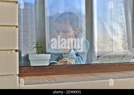 Ein trauriges Kind am Fenster in Hauskleidung ist gelangweilt, wenn es eine einsame Blume in einem Topf anschaut. Stockfoto