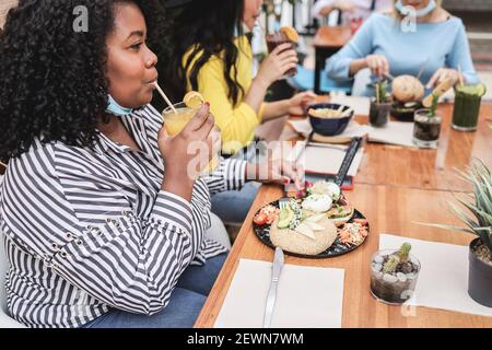 Junge multirassische Freunde beim Frühstück im Restaurant im Freien mit Masken Under Chins - Fokus auf afrikanisches Mädchen Stockfoto