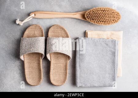 Umweltfreundliche wiederverwendbare Spa-Accessoires. Bambus-Hausschuhe, Anti-Cellulite-Massage-Bürste und Baumwollhandtücher auf grauem Stein Hintergrund Stockfoto