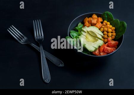 Avocado Poke Schüssel mit Kirschtomaten, Kichererbsen, Süßkartoffeln, Spinat und Lammsalat auf schwarzem dunklen Hintergrund.Nahaufnahme des vegetarischen Buddha Stockfoto