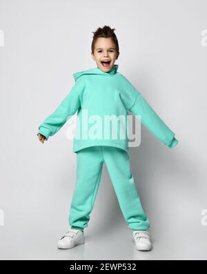 Happy Screaming Active Kid Boy in modernem Grün, mintfarbener Sportswear Anzug Kapuzenpullover und Hose steht und hält die Hände breit Stockfoto