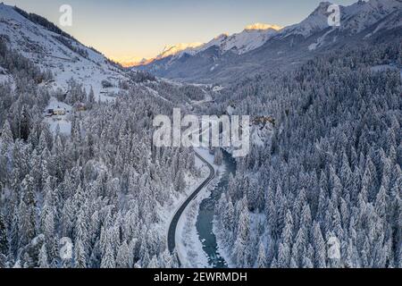 Sonnenaufgang über kurvenreicher Bergstraße und gefrorenem Fluss im schneebedeckten Wald, Zernez, Graubünden Kanton, Schweiz, Europa Stockfoto