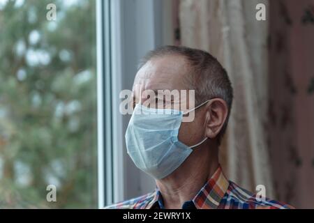 Älterer Mann mit Maske aus Covid 19 steht am Fenster. Selbstisolationsmodus. Stockfoto