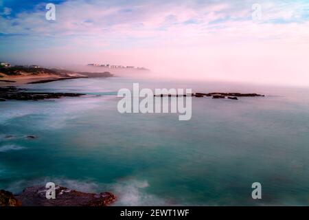 Der Nebel und Nebel, der vom Meer hereinrollt, umhüllen das Dorf Arniston. Eine lange Exposition hat das Meer und die Reflexionen von rosa Wolken auf t geglättet Stockfoto