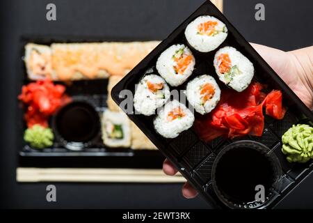 Hand hält Set Sushi in Kunststoff-Box Nahaufnahme, auf schwarzem Hintergrund. Stockfoto
