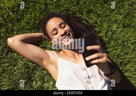 Lächelnde afroamerikanische Frau, die auf Gras liegt und eine benutzt Smartphone im Park Stockfoto