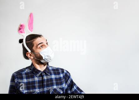 Charmante junge kaukasische Mann in medizinischen Schutzmaske und mit rosa Kaninchenohren sieht weg mit Überraschung und Bewunderung. Kreative Ostern Hintergrund Stockfoto