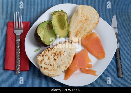 Zwei Naan-Brote, geröstet mit Lachs und halber Avocado auf weißem Teller, rote Serviette, Gabel und Messer, auf blauer Tischmatte Stockfoto