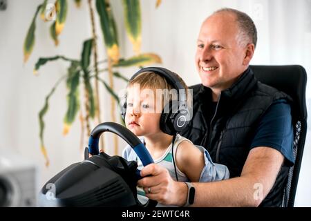 Drei Jahre alter Junge sorgfältig beobachten seinen Vater spielen Rennen Auto-Spiel auf dem Computer mit einem Lenkrad Stockfoto