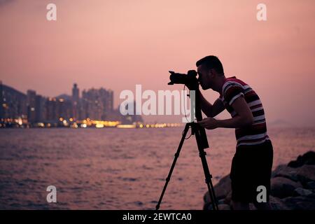 Silhouette des Fotografen mit Stativ. Junger Mann fotografiert die Skyline der Stadt. Hongkong in der Abenddämmerung. Stockfoto