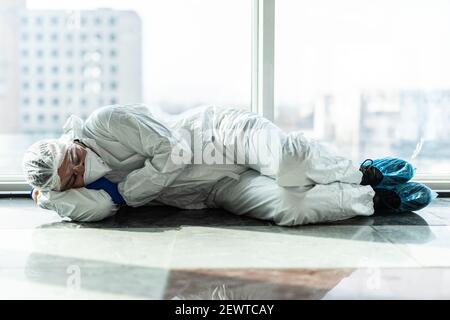 Müde asiatische Arzt in PSA Anzug sitzen und schlafen auf dem Boden, überarbeiteter Arzt Stockfoto