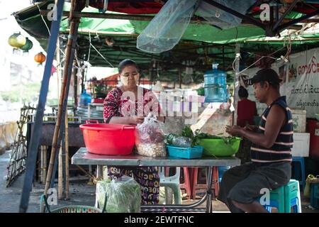 YANGON, MYANMAR - JANUAR 1 2020: Eine burmesische Frau in traditionellem Longyi und Thanaka bereitet Gemüse an einem Imbissstand mit einem burmesischen Mann zu Stockfoto