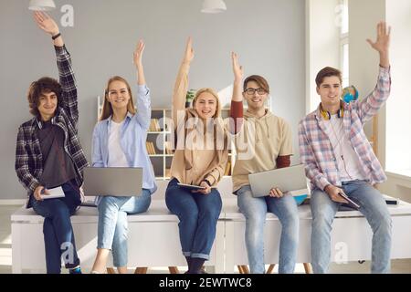 Gruppe von glücklich aktiven Schule oder College-Studenten sitzen auf Schreibtisch und Hände heben Stockfoto