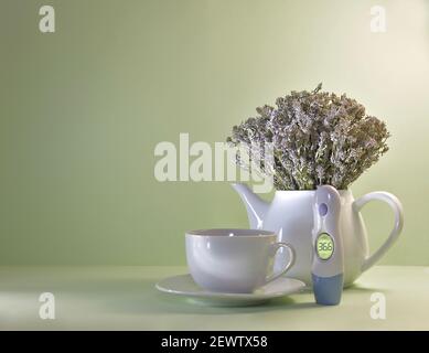 Eine Teekanne mit einem Bouquet von getrocknetem Thymian, daneben eine Tasse und Untertasse und ein Thermometer mit einem leuchtenden Bildschirm. Auf hellgrünem Hintergrund. Stockfoto