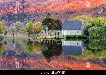 Sonnenaufgang beleuchtet die Berge in herbstlichen Farben hinter St Finbarr's Oratory und spiegelt sich im See von Gougane Barra, West Cork, Irland. Stockfoto