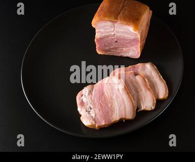 Essen, ein Stück Speck und in Scheiben geschnitten auf einem schwarzen Teller auf schwarzem Hintergrund Stockfoto