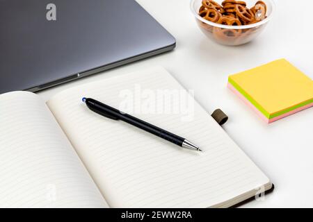 Moderner weißer Büroschreibtisch mit offenem leerem Notizbuch, Stift und anderen Hilfsmitteln mit Schale mit Snacks. Selektiver Fokus. Stockfoto