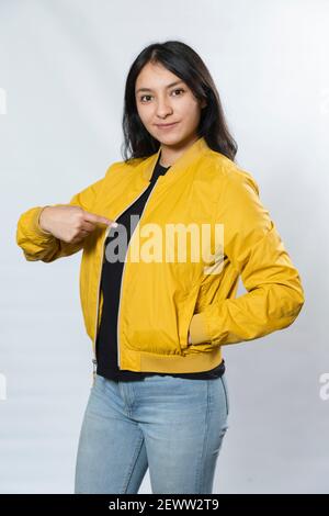 Mockup women's yellow bomber jacket-Portrait of young Hispanic woman pointing at her jacket on white background Stockfoto