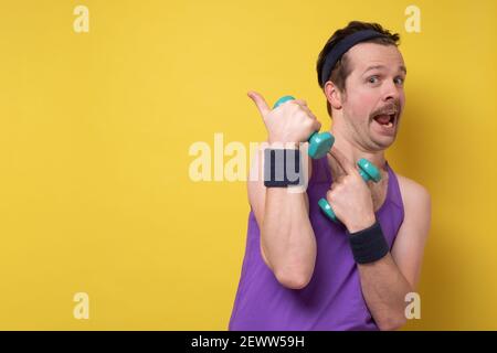 Junger lustiger kaukasischer Mann mit Hanteln, die lächeln und zur Seite zeigen, zeigt etwas auf leeren Raum. Stockfoto