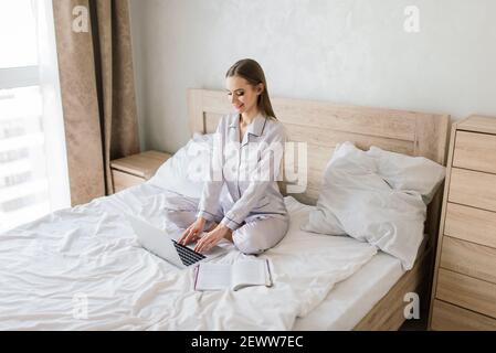 Foto der jungen fröhliche Frau in Pyjama Tippen auf Laptop und lächelnd beim Sitzen auf dem Bett im Zimmer Stockfoto