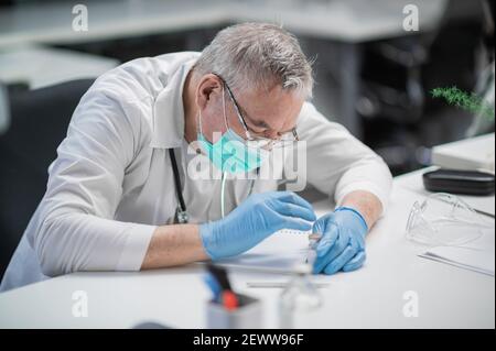 Ein Arzt mit einer Brille und einer medizinischen Maske nimmt Aufzeichnungen auf Informationen über Patienten in seiner Klinik Stockfoto