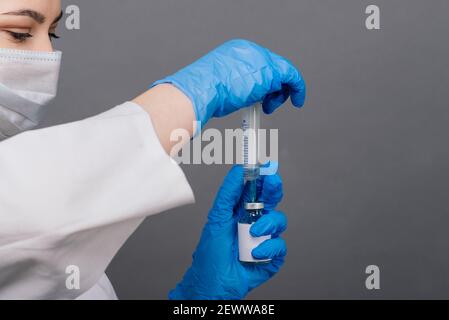 Seriöse Ärztin in einer medizinischen Maske hält Spritze und Impfstoff isoliert auf grau Stockfoto