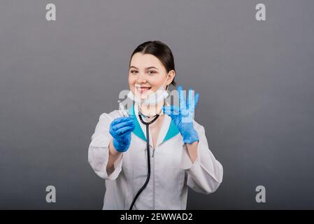 Seriöse Ärztin in einer medizinischen Maske hält Spritze und Impfstoff isoliert auf grau Stockfoto