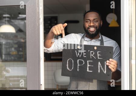 Konzept für kleine Unternehmen. Lächelnder afroamerikanischer Typ Backwaren in einer Schürze zeigt Finger auf ein offenes Schild in seiner Hand. Ein multirassischer Cafe-Besitzer lädt zum Besuch wiedereröffneter Lage ein Stockfoto