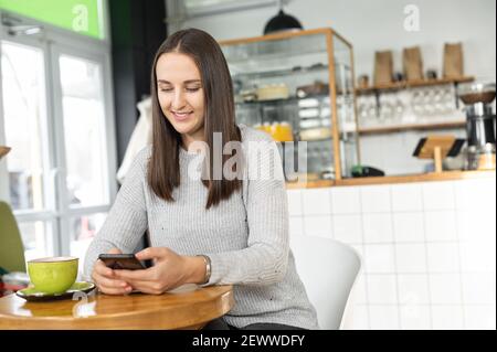 Lächelnde junge Brünette Frau genießt einen Morgenkaffee im gemütlichen Café sitzend und auf dem Smartphone chattet, hat Studentin eine Pause mit Telefon und Heißgetränk in der Cafeteria Stockfoto